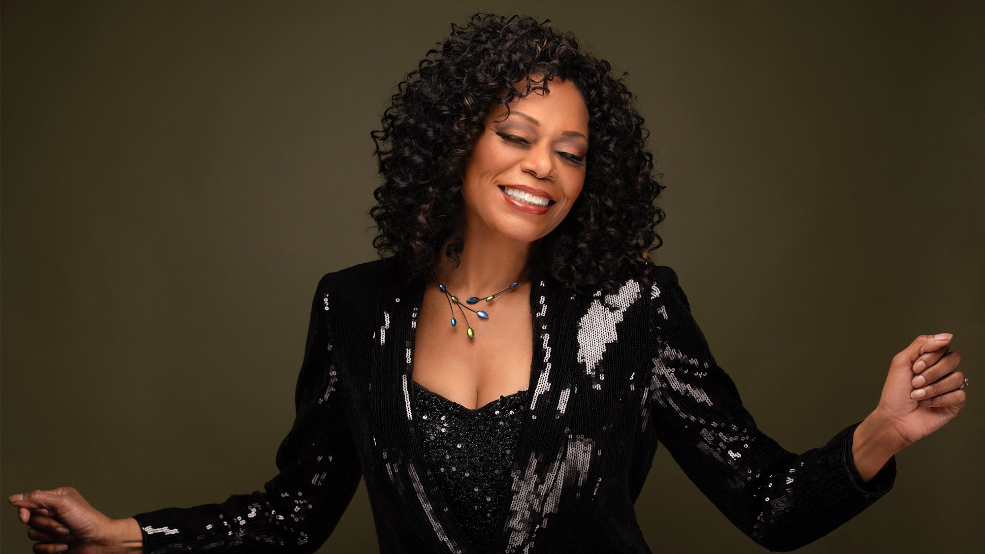 Stephanie Pope beams with closed eyes, wearing a sequined black jacket and beaded necklace. Channeling Livin' It Up energy, she poses joyfully with both arms bent against an olive green background.