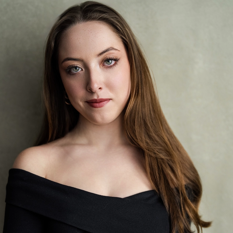 Maggie Stephens, a young woman with long brown hair and fair skin, looks at the camera in a black off-the-shoulder top. She stands against a neutral, softly textured background, wearing natural makeup and a slight, confident smile.