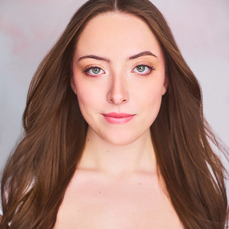 Maggie Stephens, a young woman with long brown hair and green eyes, looks directly at the camera with a neutral expression. She has fair skin, subtle makeup, and her hair flows over her shoulders against a soft, light background.