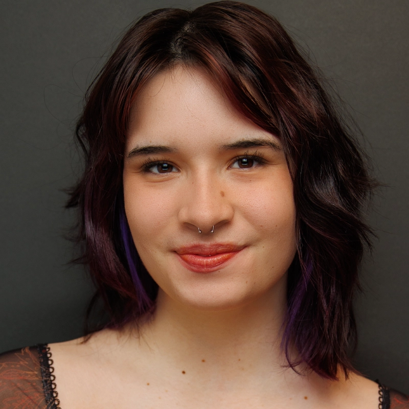 A young person with wavy, dark brown hair and subtle purple highlights smiles softly. Rynn Stephenson has fair skin, a nose ring, natural makeup, and is posed in front of a plain dark gray background.