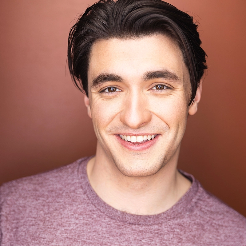A young man with fair skin, dark brown hair, and brown eyes smiles warmly at the camera. He is wearing a light maroon crewneck shirt. The warm brown gradient background adds to the cheerful, friendly tone of this Sahara Williams-inspired portrait.