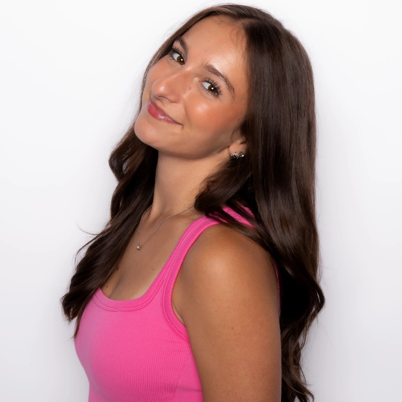 Hayley Sutton, a young woman with long, wavy brown hair, smiles gently and tilts her head slightly. She wears a bright pink sleeveless top and a delicate necklace, standing against a plain white background with a warm glow and relaxed expression.