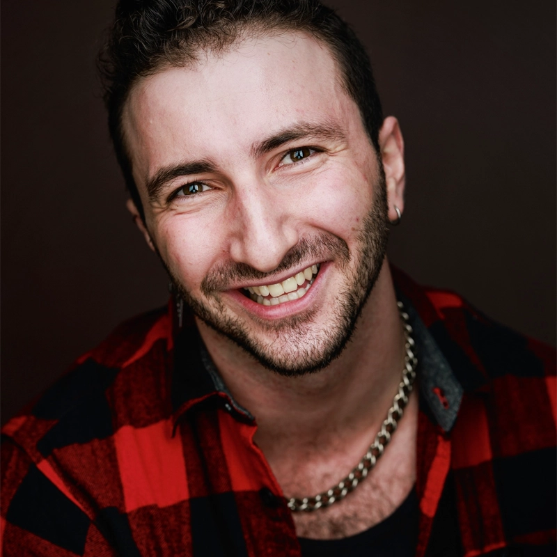 A smiling man with short curly hair and a trimmed beard, identified as Jared Svoboda, wears a red and black plaid shirt and a thick silver chain necklace. He has a nose ring in his left nostril and is photographed against a dark background.