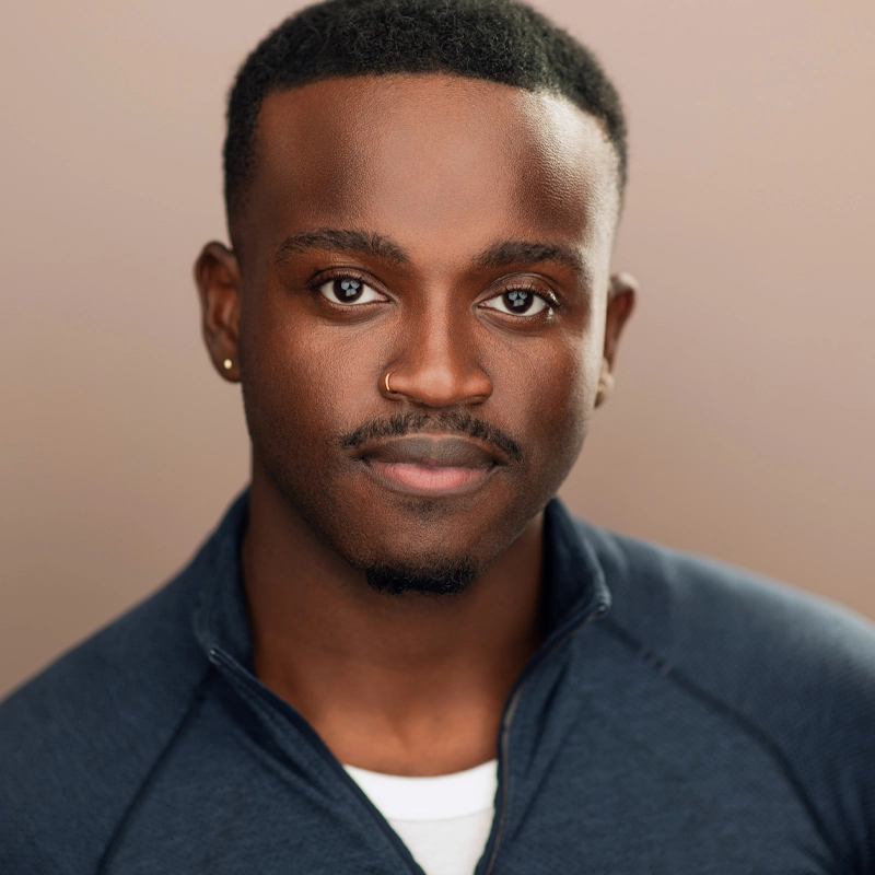Michael Swain-Smith, a young Black man with short hair, gazes softly at the camera. He wears a navy zip-up jacket over a white shirt, with a small hoop earring and a nose ring. The soft beige background adds a warm, professional touch.