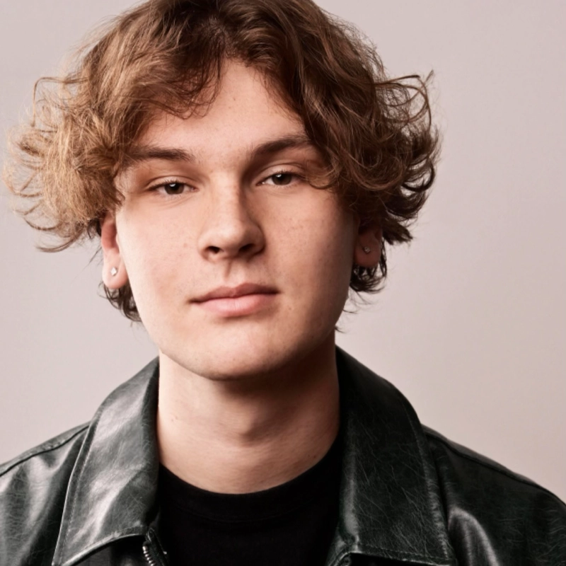 A young person with wavy, light brown hair, wearing small hoop earrings, a black shirt, and a dark leather jacket, faces the camera with a neutral expression against a light gray background—resembling Daniel Swanson.