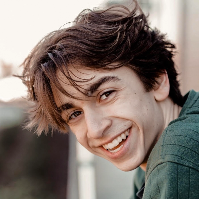 A young person with tousled brown hair smiles broadly, showing teeth. They wear a green top and are outdoors in soft, natural light, with a blurred background for a warm, candid portrait style.