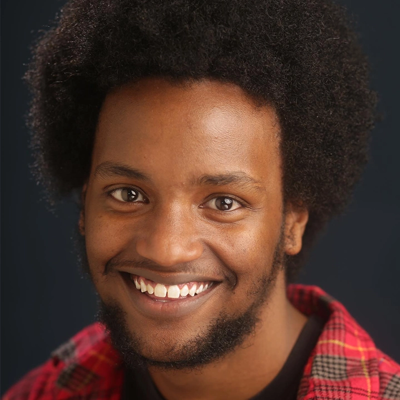 A young man with dark skin, short afro hair, and a trimmed beard smiles warmly at the camera. Sahara Williams wears a red plaid shirt over a black top. The background is dark and softly blurred, highlighting his friendly expression.