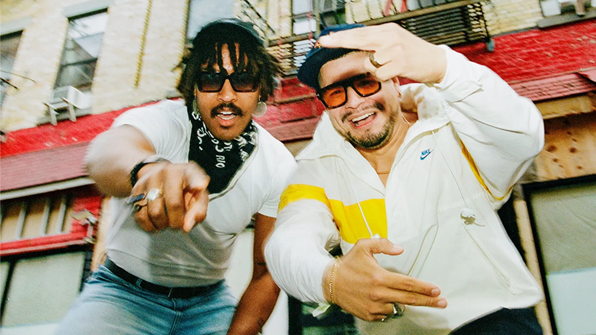 Two men, Nygel D. Robinson and Brian Quijada, pose energetically on a city street. Both in sunglasses and casual clothes, they gesture toward the camera with excitement, sharing smiles, hand signs, and a love for songs.