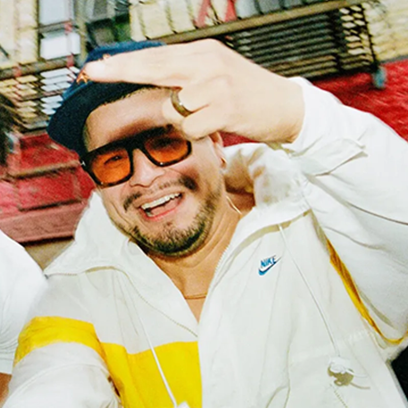 A man wearing a white and yellow Nike jacket, a blue cap, and large sunglasses smiles while making a rude middle finger gesture toward the camera. He has a beard, mustache, and a visible gold ring. The background features red and yellow walls and metal bars.