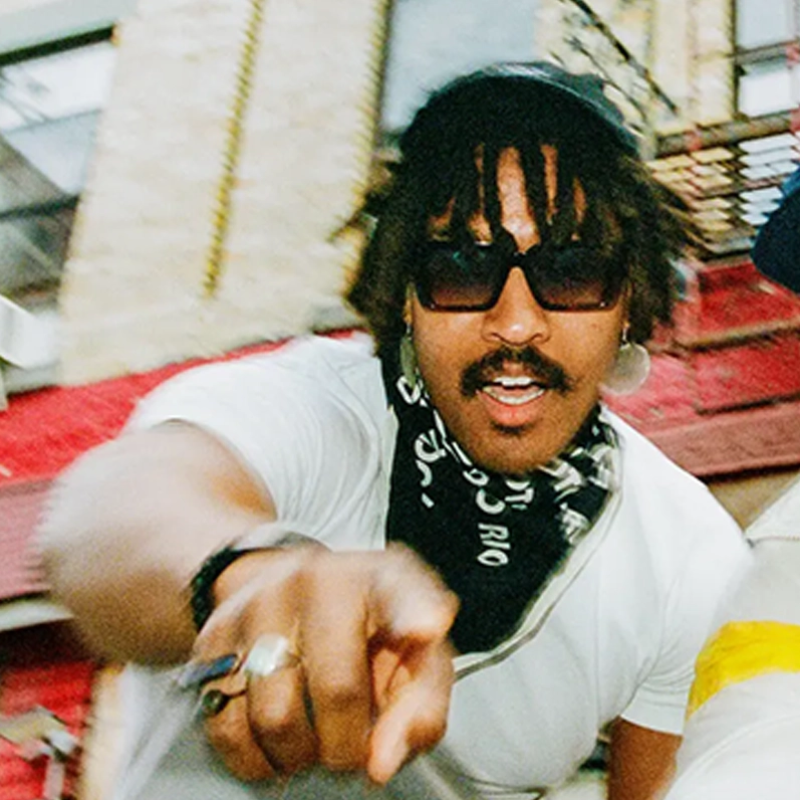 A man with medium brown skin, dreadlocks, a mustache, and sunglasses wears a white t-shirt and dark bandana around his neck. He points toward the camera, outdoors with urban buildings in the blurred background.