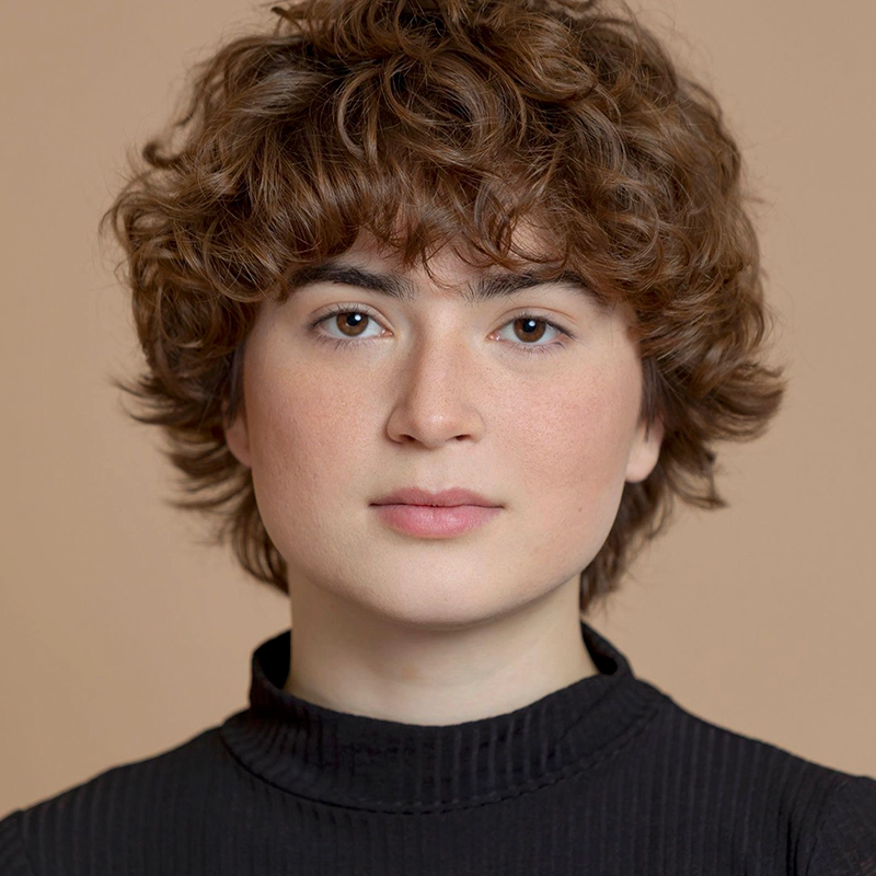 A person with short, curly brown hair and fair skin looks directly at the camera with a neutral expression. They are wearing a black, ribbed high-neck top. The background is a solid light beige color.