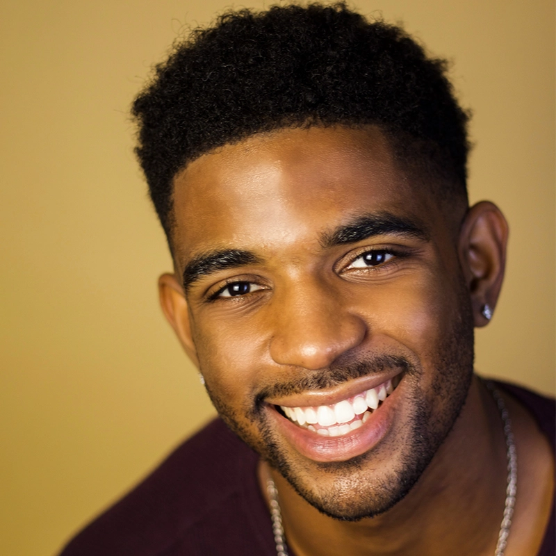 A young man with short curly hair smiles warmly at the camera, showing his teeth. RJ Vandenbrouck has a trimmed beard, small hoop earrings, and wears a maroon shirt with a silver chain against a light mustard yellow background.