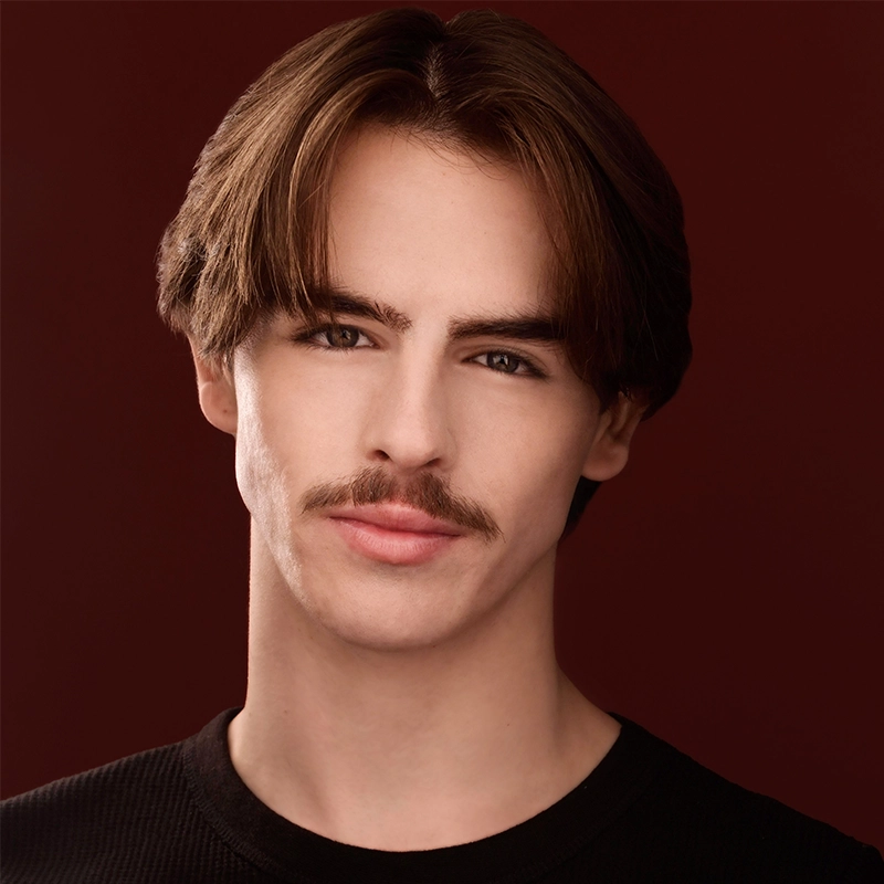 A young man with fair skin, straight brown hair parted in the middle, and a thin mustache. Rynn Stephenson is wearing a black crew-neck shirt against a dark red background, looking directly at the camera with a slight smile.