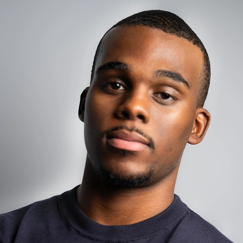 A young man with short hair and a trimmed goatee looks at the camera with a neutral expression. He is wearing a dark crew-neck sweater. The plain, softly lit background echoes the minimalist style found in Kenzie Woolridge’s portraits.