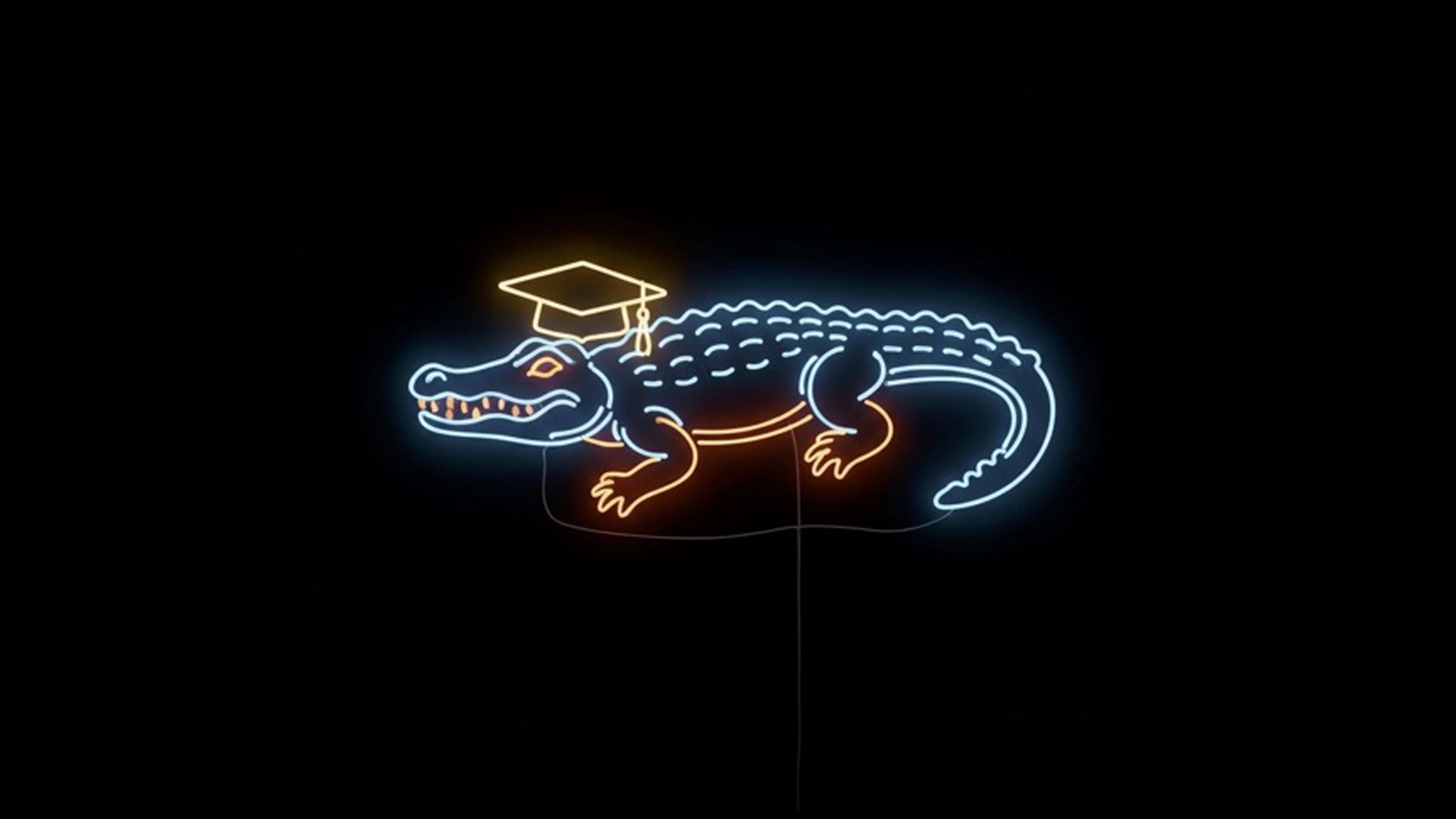 A bright neon sign outlines a smiling alligator wearing a graduation cap, celebrating the University of Florida BFA Musical Theatre Class of 2026. The glowing blue gator features orange accents and a yellow-capped head against a black background.