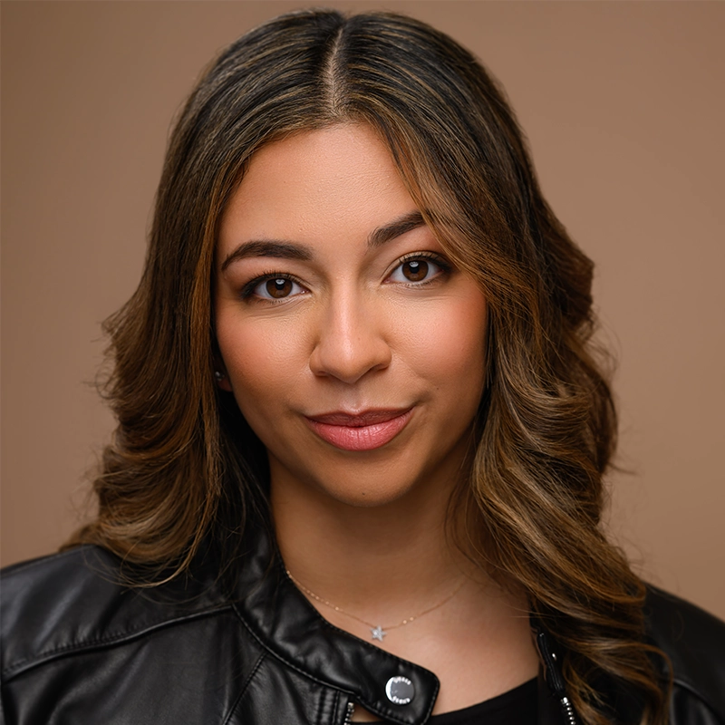 A woman with wavy, light brown hair and medium skin tone looks directly at the camera. She wears a black leather jacket, a delicate necklace with a small star pendant, and subtle makeup. The background is a solid light brown color.