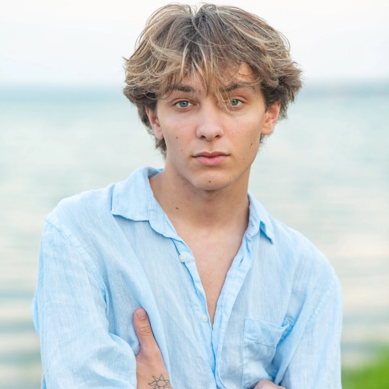 A young person with tousled, light brown hair and green eyes stands outdoors by water. Wearing a light blue, unbuttoned shirt, they have a tattoo on their forearm—reminiscent of Charles Van Flaherty's relaxed style. The background is softly blurred.