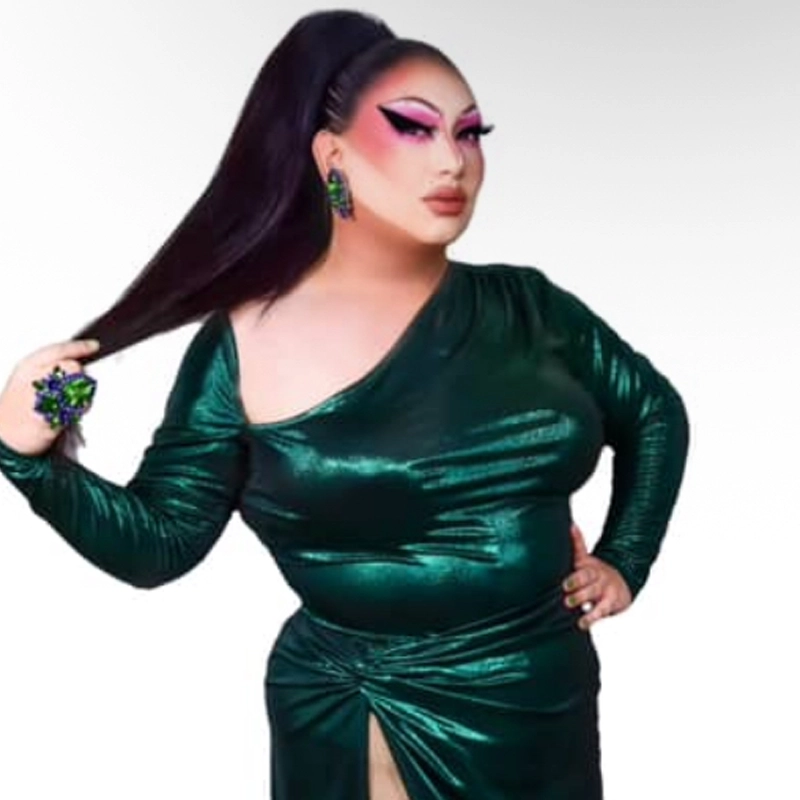A person wearing a shiny green Versace one-shoulder gown poses with one hand on their hip and the other holding their long, high ponytail. They have bold pink eyeshadow, dramatic eyeliner, sculpted brows, and large green earrings and ring. The background is white.