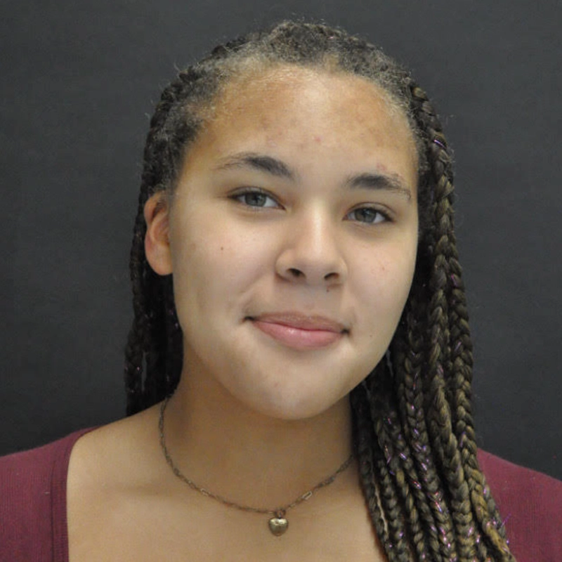 Peyton Amaral, a young woman with long, braided hair, a light brown complexion, and hazel eyes, smiles slightly. She wears a maroon top and a delicate heart pendant necklace, standing out against the plain, dark background.