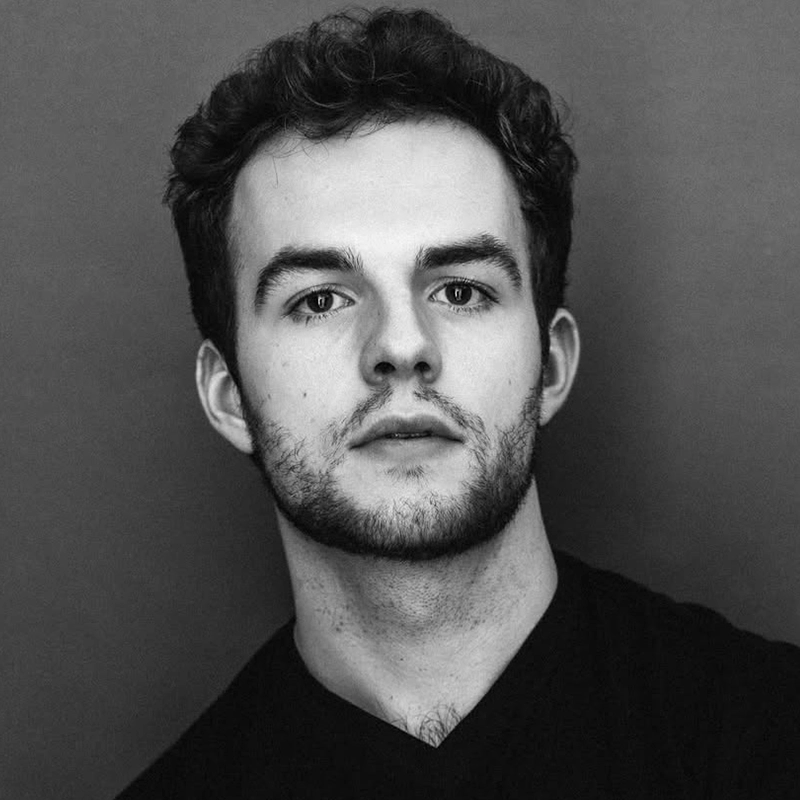 A young man with short, curly dark hair and a trimmed beard looks directly at the camera. He has light skin, expressive dark eyebrows, and is wearing a dark V-neck shirt. The background is a plain, solid color in a neutral tone.