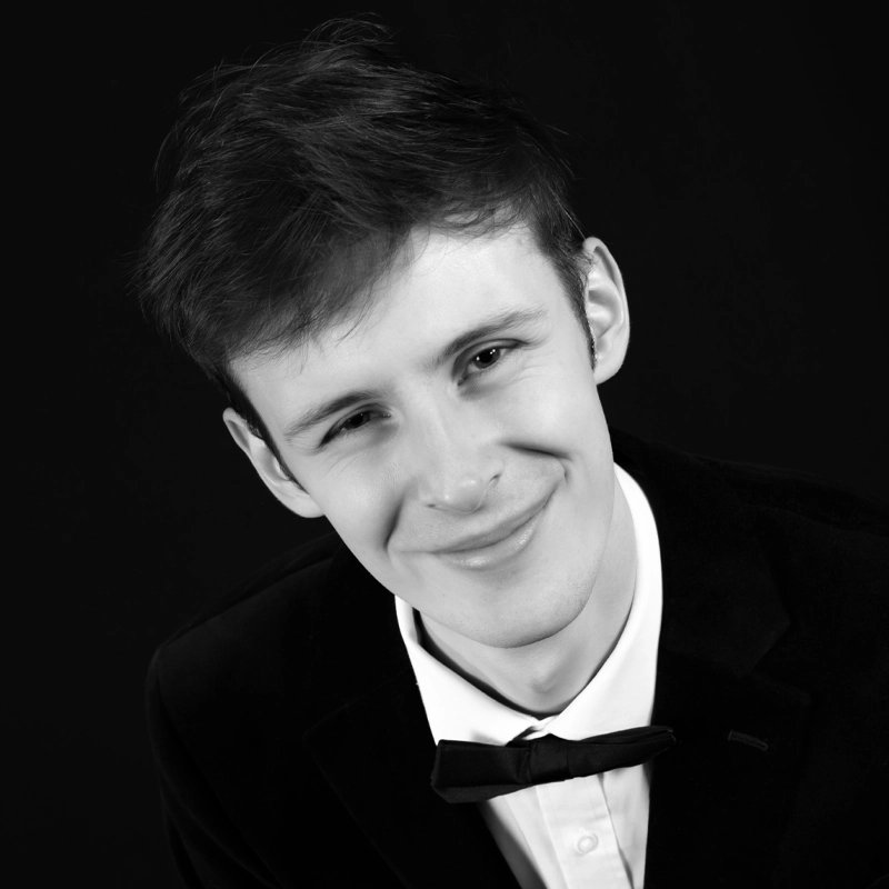 A young man with short, dark hair smiles gently at the camera. Ricky Westrip is dressed in a black tuxedo jacket, white dress shirt, and black bow tie. The solid black background gives the portrait a formal and elegant look.