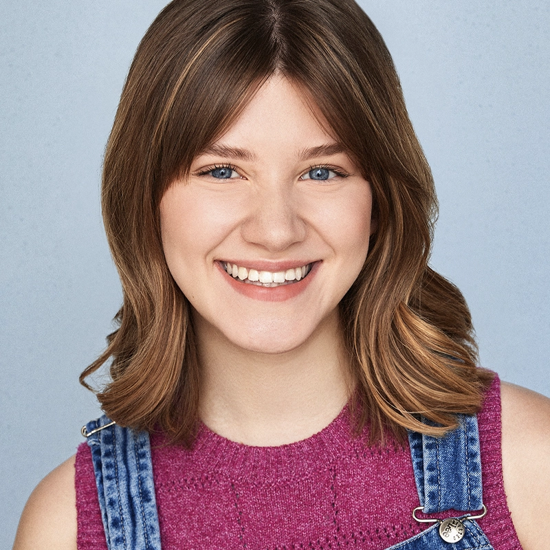 A young woman with shoulder-length brown hair smiles warmly at the camera. She has blue eyes and wears a sleeveless magenta knit top with blue denim overalls. The background is a soft, light blue, creating a friendly and approachable atmosphere.