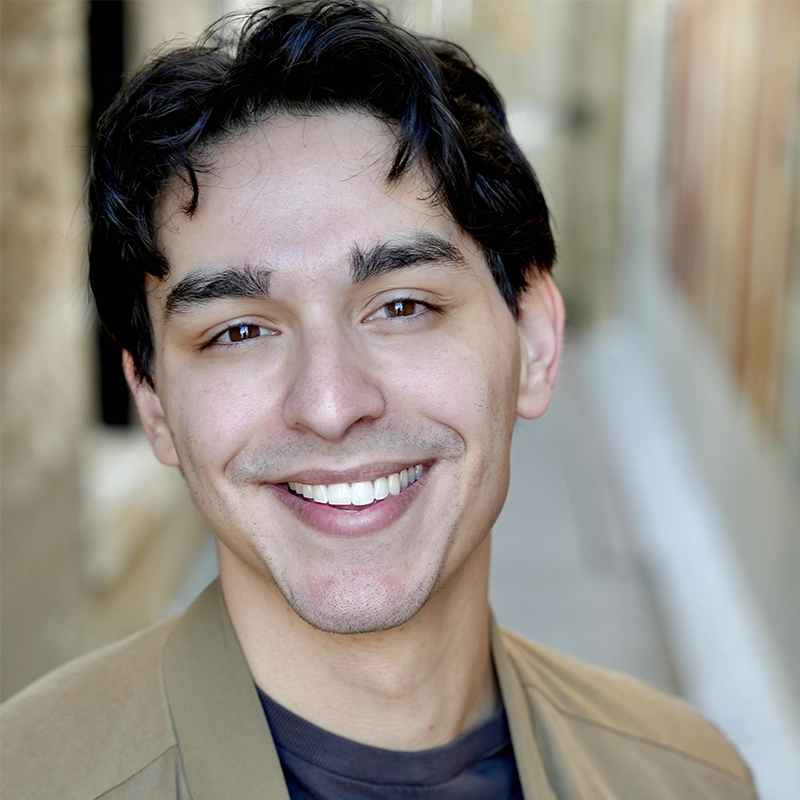 A young man with wavy dark hair and light skin smiles directly at the camera. He is wearing a tan jacket over a dark shirt. The background is softly blurred, suggesting an outdoor urban setting with stone walls and an alleyway.