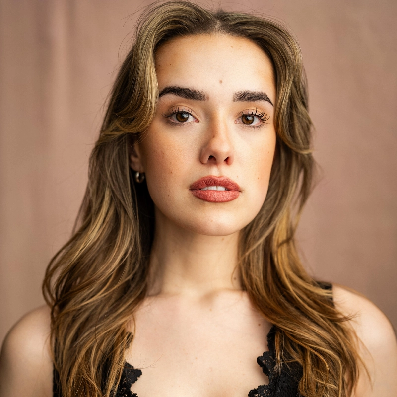 Audrey Williams, a young woman with long, wavy brown hair and light skin, faces the camera. She wears natural makeup, a neutral lip color, and a black lace top. The soft, light brown background highlights her calm, confident presence.