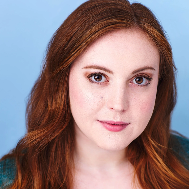 A young woman with long, wavy auburn hair and fair skin looks directly at the camera. She has brown eyes, lightly arched brows, and soft pink lips. She wears natural makeup and a green top, with a light blue background behind her.