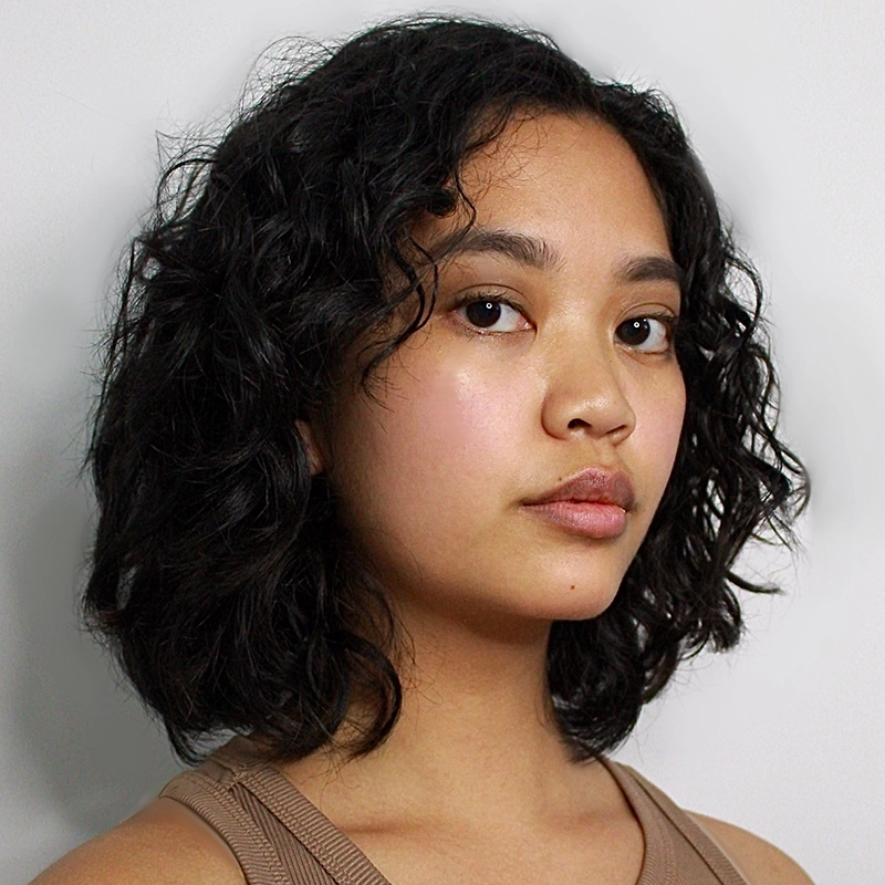 A young woman with medium skin, dark wavy shoulder-length hair, and natural makeup looks confidently at the camera. Caroline Weston wears a sleeveless beige top and stands against a plain white background.