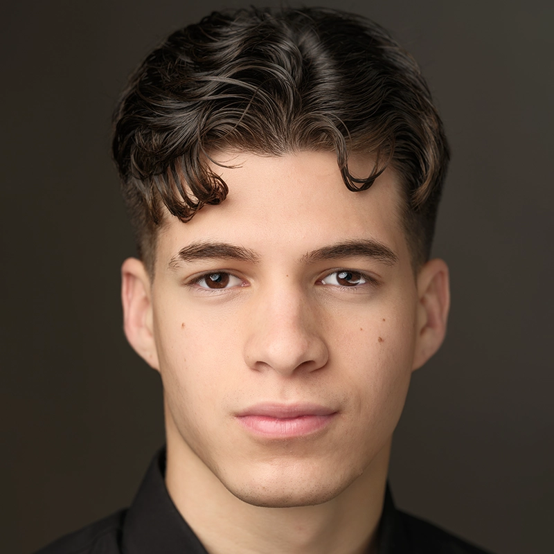 Young man with short, wavy dark hair styled back, and two curls falling on his forehead. He has fair skin, brown eyes, and a neutral expression. His look is reminiscent of a Caroline Bowers portrait. He wears a black shirt; the background is softly blurred.