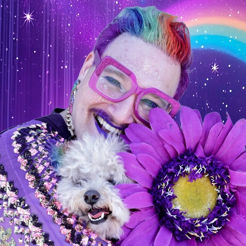 Caroline Weston, a joyful person with rainbow hair, bright purple glasses, and a sequined outfit, smiles while holding a fluffy white dog and a large purple flower against a cosmic backdrop of stars, sparkles, and a vibrant rainbow.