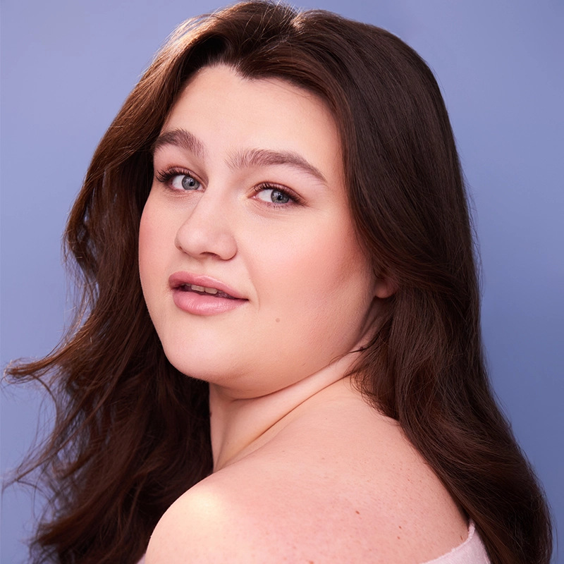 Rynn Stephenson, a woman with long brown hair and fair skin, looks over her shoulder at the camera, smiling softly. She has natural makeup with light pink lipstick and is posed against a plain, light blue background.