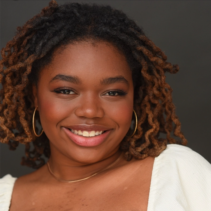 Kenzie Woolridge, a young woman with medium brown skin and short, curly hair, smiles warmly in gold hoop earrings and a thin gold necklace. She is dressed in a white top and posed against a plain, dark background.