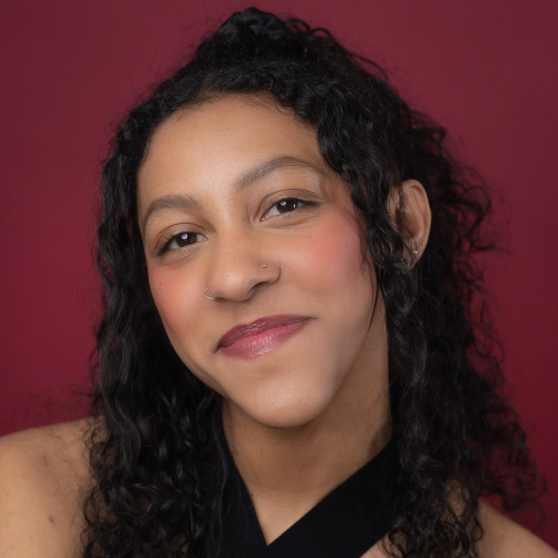 Sabrea Aijalon, a young woman with curly black hair and a nose ring, smiles softly. She wears natural makeup with pink blush and lipstick, a black top, and stands against a deep red background for a warm, inviting feel.