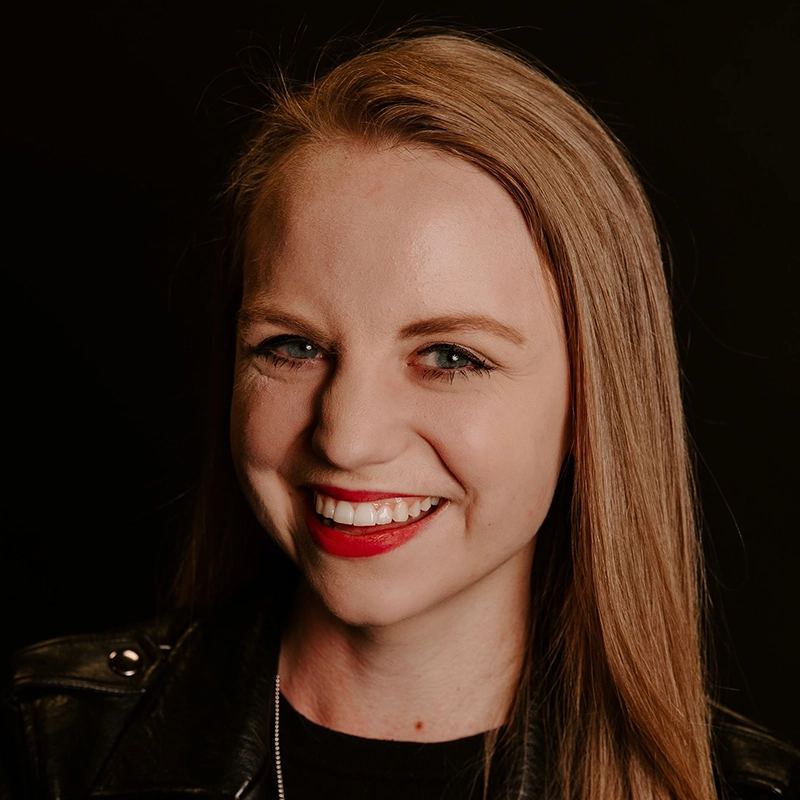 Isabel Marin Young, with straight blonde hair, smiles warmly at the camera. She wears a black leather jacket and red lipstick. The dark background makes her face and hair stand out brightly.