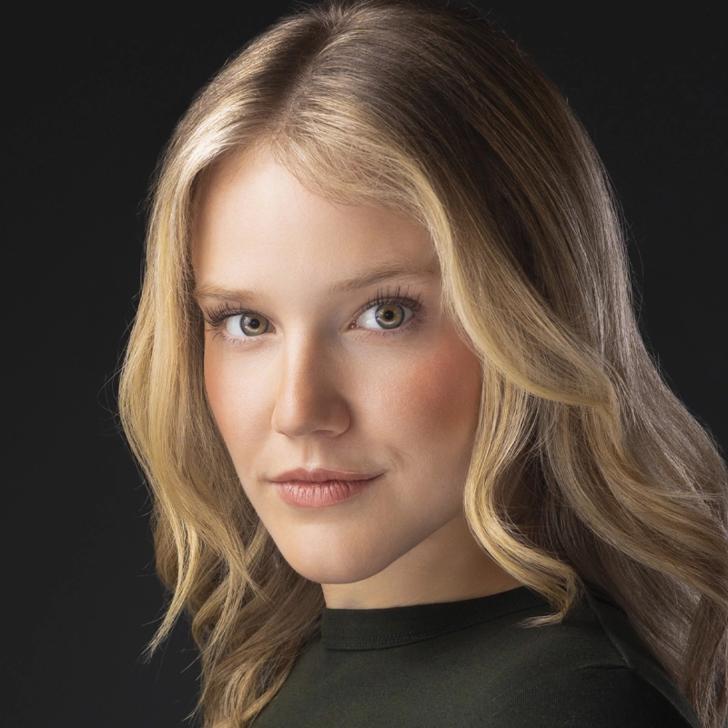 A young woman, Beverly Anderson, with long, wavy blonde hair and fair skin looks confidently at the camera. She has hazel eyes, subtle makeup, and wears a dark green top. The dark background keeps focus on her face.