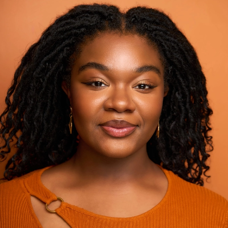 Blessett Anderson, a woman with long black curly hair, smiles gently at the camera. She wears gold earrings and an orange top with a decorative ring on the shoulder. The warm lighting and solid orange background complement her look perfectly.