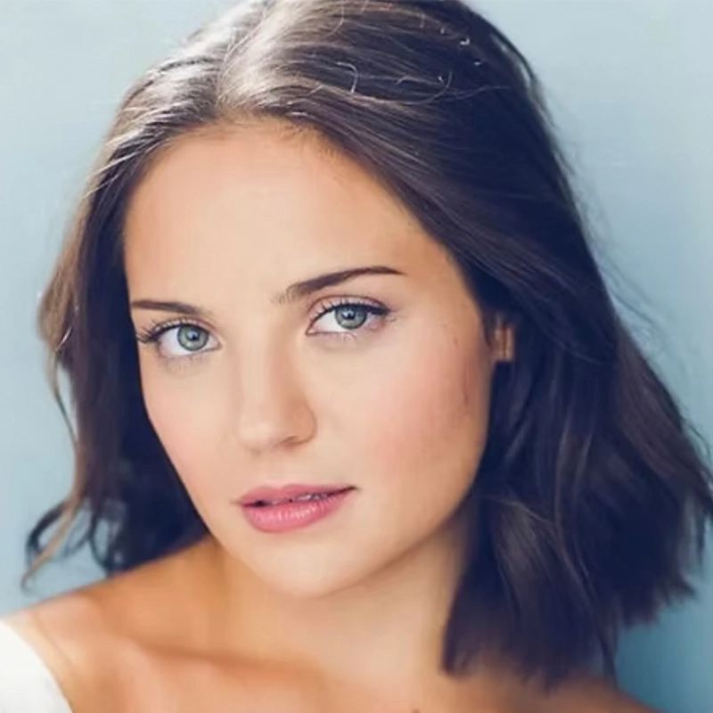 A young woman with fair skin, blue eyes, and shoulder-length brown hair looks at the camera. She has a slight, natural smile and wears light makeup. The background is a soft blue, and her outfit is off-white and off the shoulder.