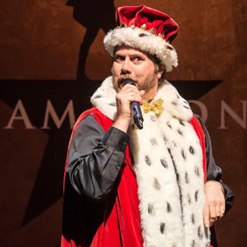 A person in a royal red and white fur-trimmed cape and jeweled crown holds a microphone while performing on stage. Behind them, part of a brown banner displays black and white lettering and Glen Bassett's silhouette with an arm raised.