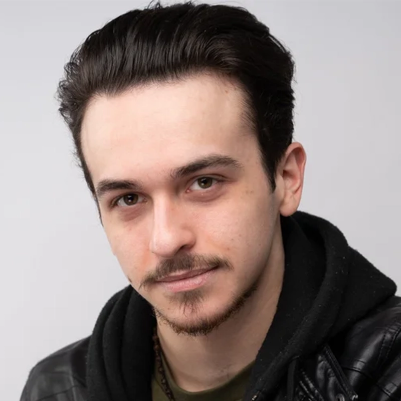 Bryce Hayden, a young man with fair skin and short dark hair, looks at the camera with a slight smile. Sporting a thin mustache and goatee, he wears a black leather jacket over a black hoodie against a plain light gray background.