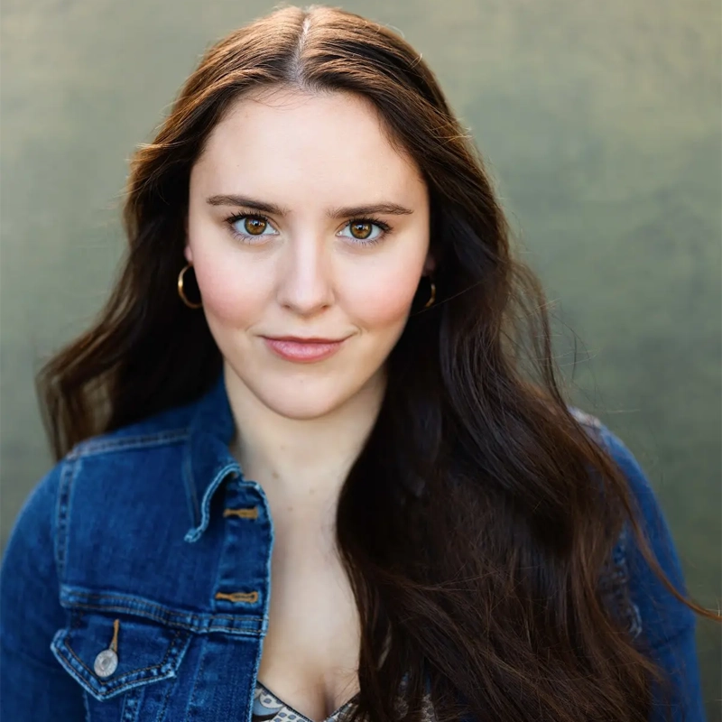 Carolyn Byrne, a young woman with long, wavy brown hair, wears a blue denim jacket over a patterned top. She faces the camera, smiling softly with neutral makeup and gold hoop earrings against a muted green, softly blurred background.
