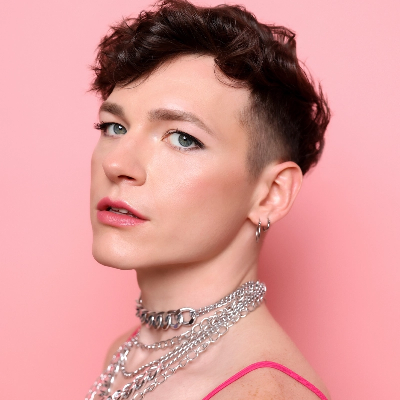 Liam Collins, with short, curly brown hair and blue eyes, poses against a pink background. They wear subtle makeup—pink lipstick—accessorized by silver chain necklaces and small hoop earrings. A thin pink strap is visible on their shoulder.