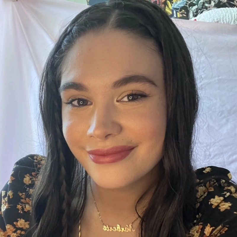 A young woman, Melanie Concepcion, with long, dark hair styled in two braids smiles at the camera. She wears a floral-patterned black top and a gold name pendant. A white sheet is draped behind her, with colorful fabrics in the background.
