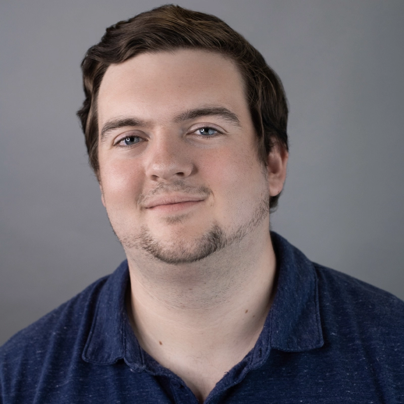 A young man with short brown hair and light skin smiles softly at the camera. He has blue eyes, light facial hair, and is wearing a dark blue collared shirt. The background is a plain, neutral gray—this is Alexander Cousins.