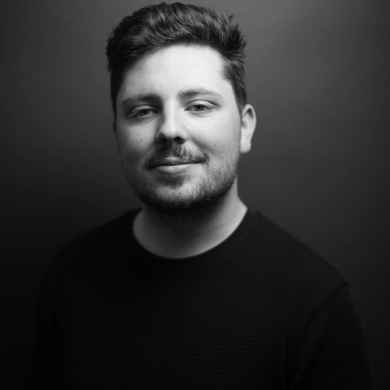 A young man, Michael Cuschieri, with short, dark hair and a trimmed beard smiles slightly while facing the camera. He wears a textured, dark long-sleeve shirt. The plain, dark background and soft lighting give the portrait a professional, studio feel.