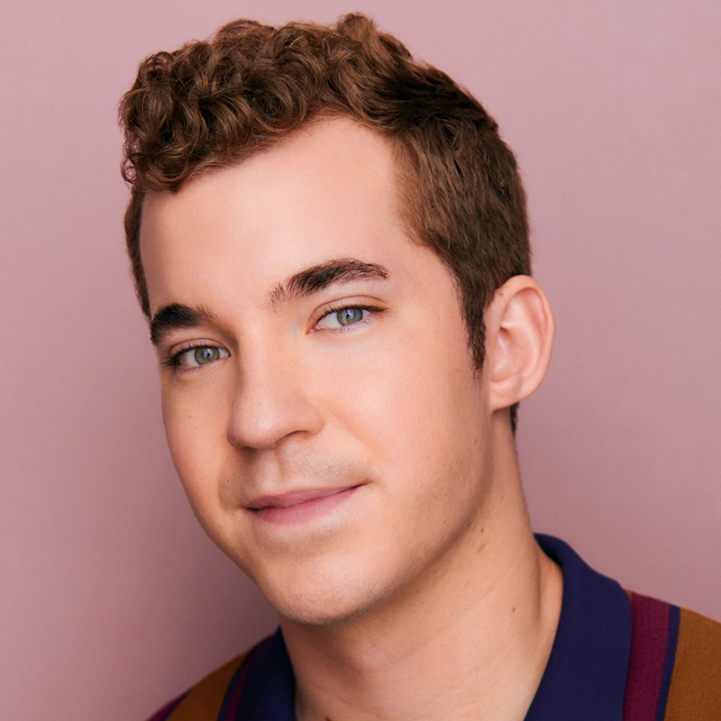 A young person with short, curly light brown hair and blue eyes smiles softly. They are wearing a collared shirt with maroon and navy stripes. The background is a solid light pink color. The lighting is soft and even.