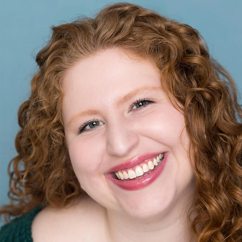 A woman with fair skin and long, curly red hair smiles warmly at the camera, reminiscent of Danielle Cohn. She has blue eyes and wears rosy lipstick. The soft blue background creates a cheerful and inviting portrait.