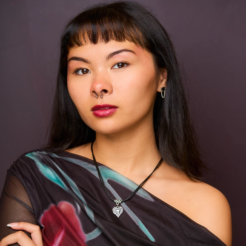 Yazmin DeJesus, with straight black hair and blunt bangs, poses against a dark background. She wears a sheer, floral-patterned top off one shoulder, a heart-shaped pendant necklace, silver earrings, and a septum ring.