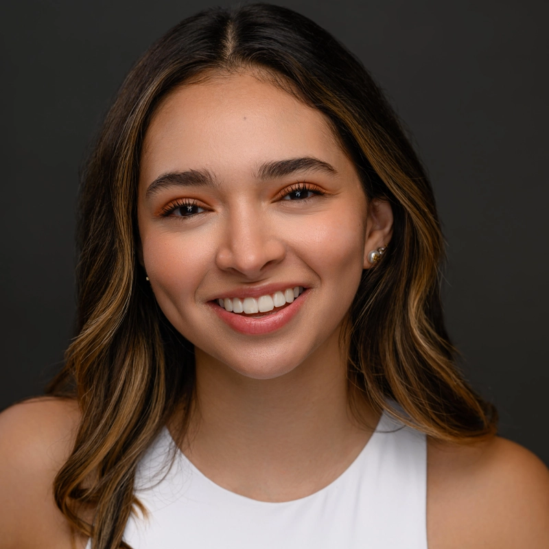 Daniela Diaz, a young woman with long brown hair in loose waves and subtle highlights, smiles warmly. She wears a white sleeveless top, small stud earrings, and natural makeup with peachy eyeshadow against a dark, plain background.